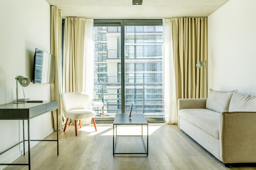 Sala de estar del Urban Studio con sofá, escritorio de trabajo, silla de diseño y gran ventanal, ideal para estadías largas.