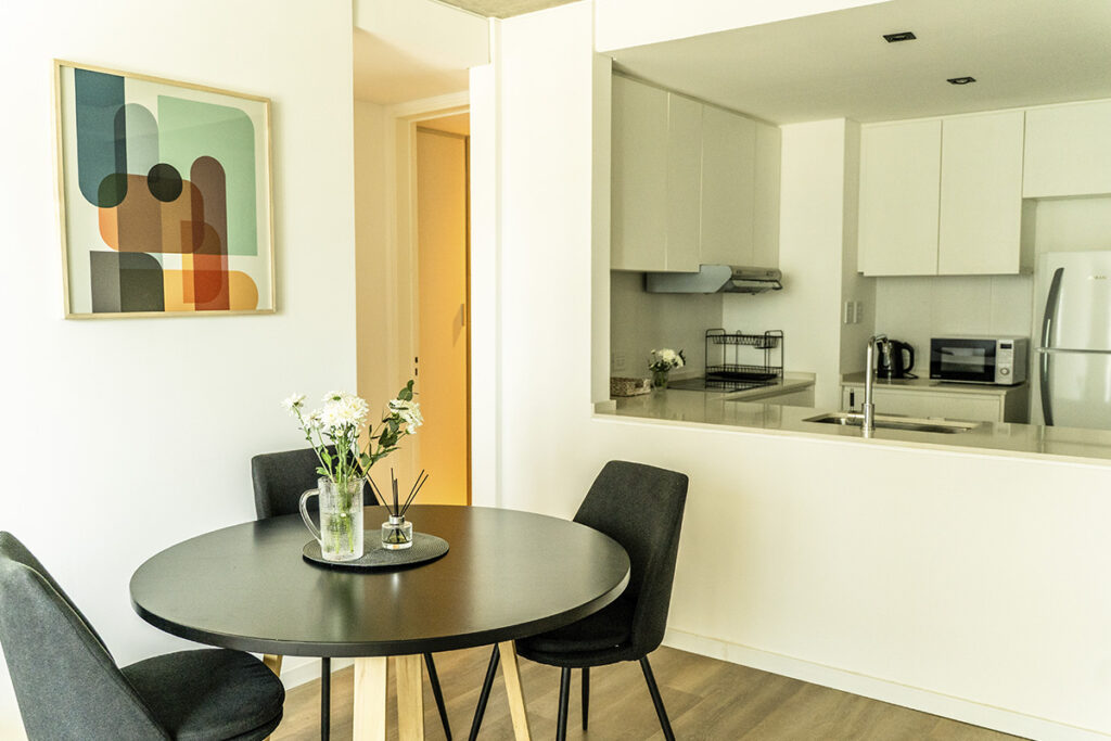 Mesa de comedor redonda negra, sillas oscuras, cuadro abstracto y vista parcial a la cocina a través de la abertura.