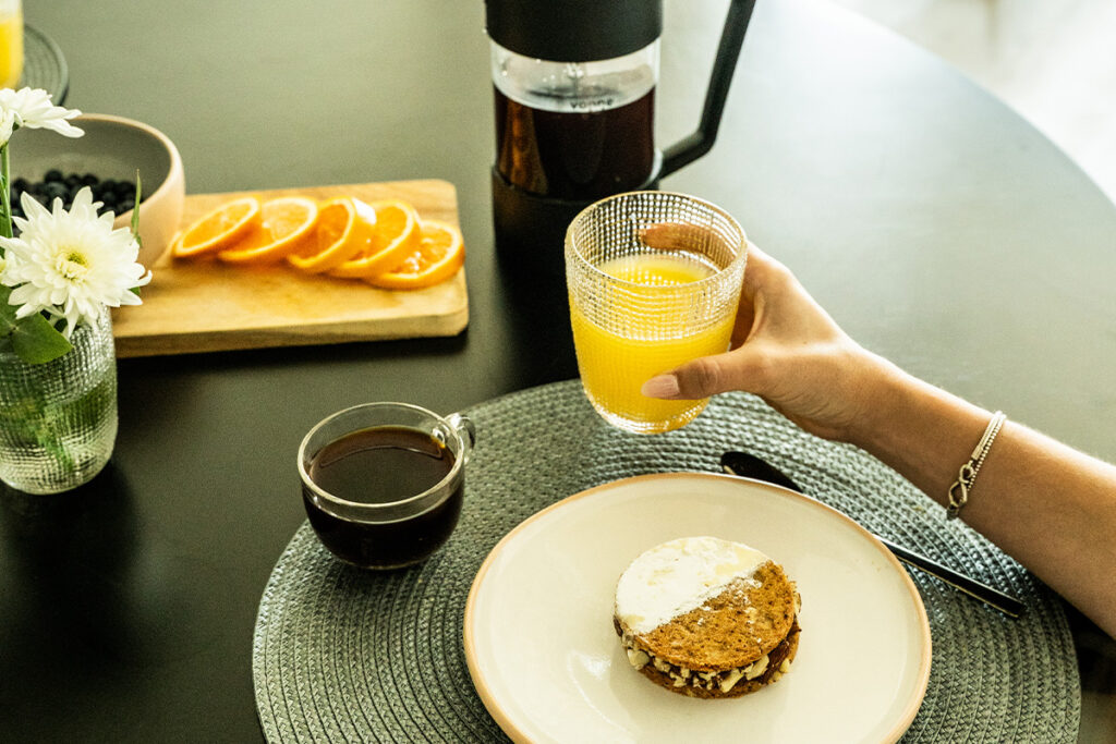 Primer plano de una mano sosteniendo un vaso de jugo de naranja, con prensa francesa de café en la mesa