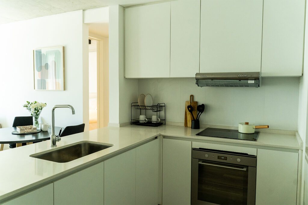 Cocina en L con mesada blanca, pileta de acero inoxidable, horno empotrado y detalles de tabla de madera.