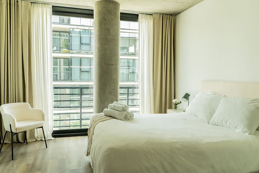 Dormitorio con cama Queen, sillón de acento blanco y pilar de hormigón a la vista junto al ventanal.