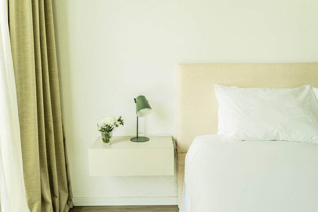 Detalle de la mesita de noche flotante con lámpara de diseño verde, flores blancas frescas y cabecera tapizada.