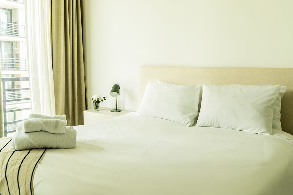 Primer plano de la cama Queen con toallas dobladas, manta a rayas y cortinas que dan al balcón.