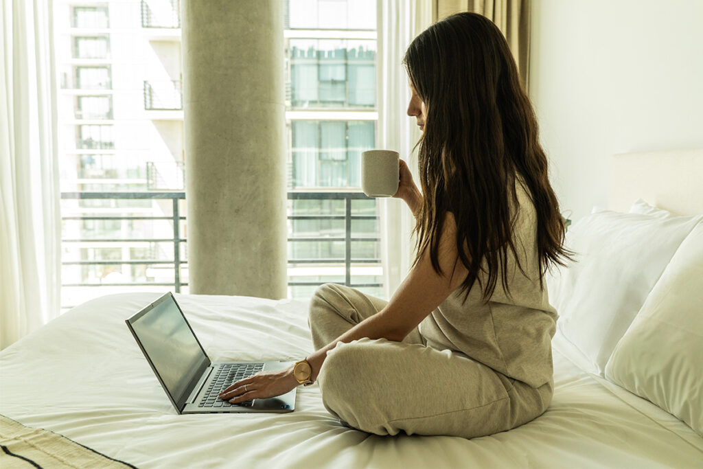 Mujer con ropa cómoda trabajando en su laptop y tomando café, sentada en la cama del estudio.