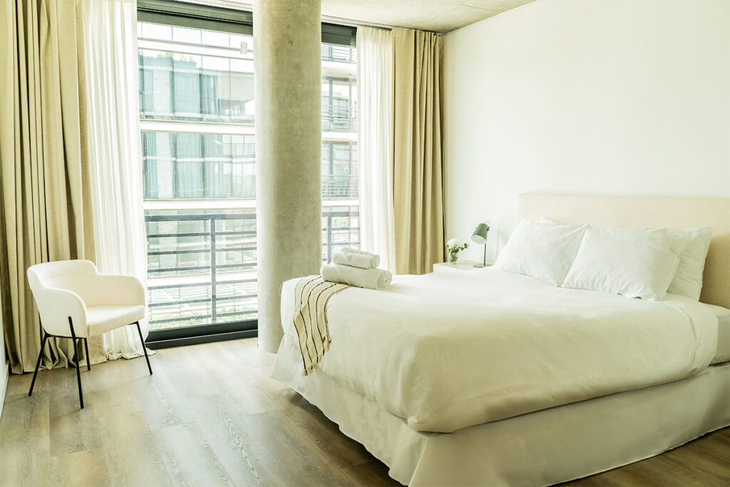 Dormitorio completo con cama, sillón de acento blanco y ventanal de piso a techo, bien iluminado.