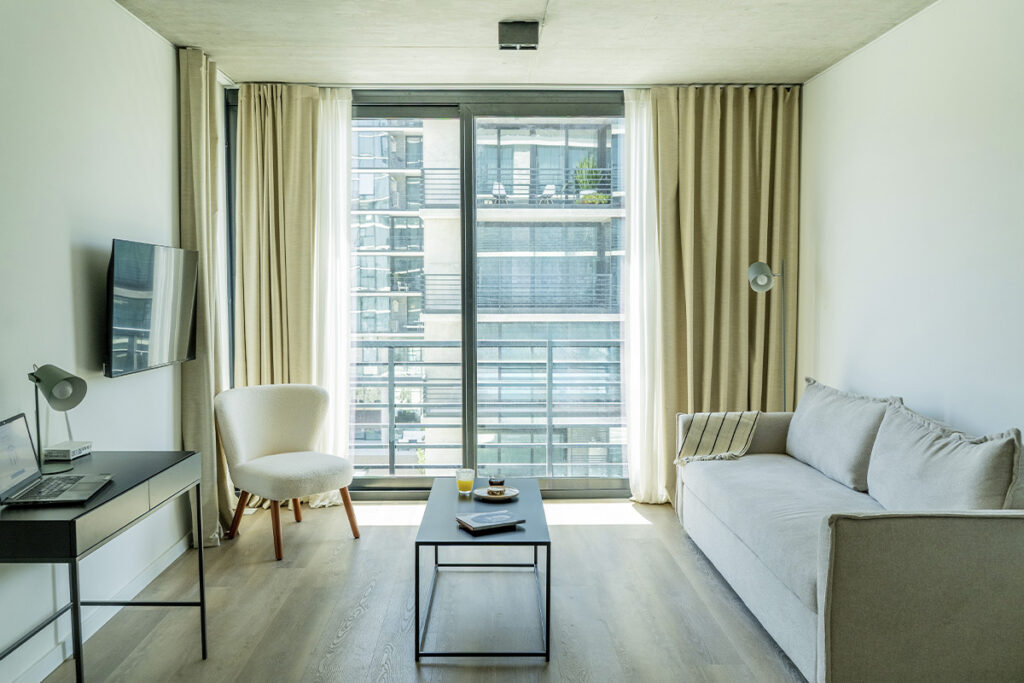 Living con escritorio de trabajo (laptop visible), silla de acento, sofá y ventanal de pared a pared.