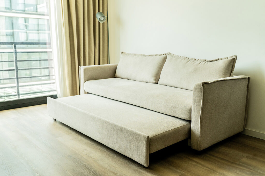 Sofá cama beige parcialmente desplegado, mostrando la opción de alojamiento adicional para un huésped.