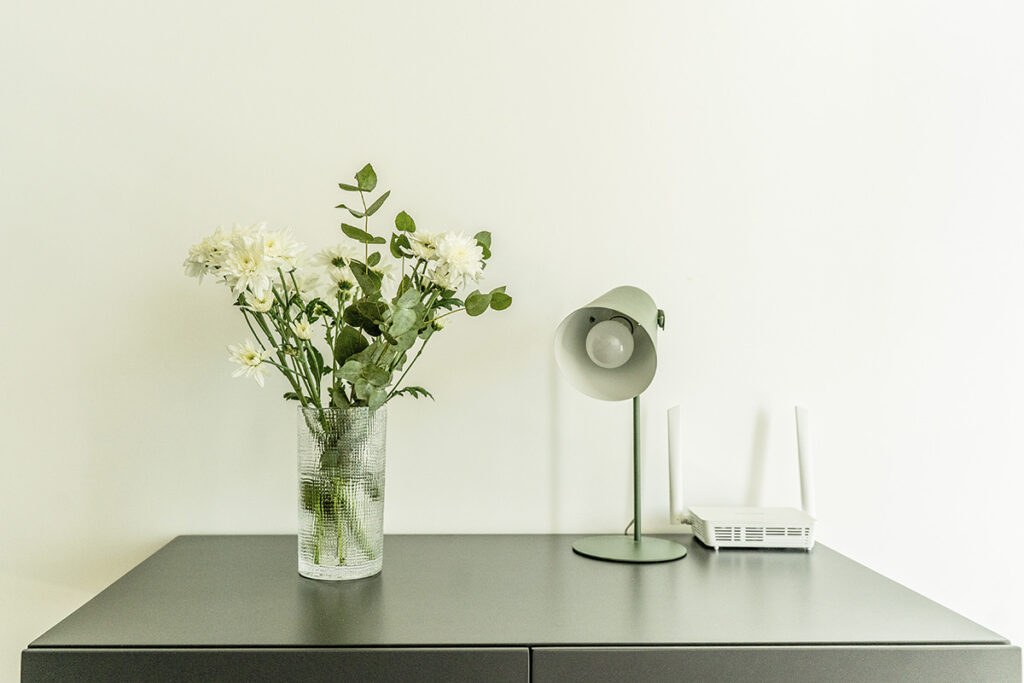 Detalle del escritorio con router WiFi, lámpara de diseño y florero con flores blancas para ambientación.