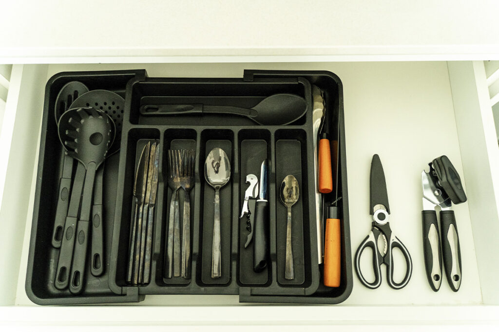 Cajón abierto mostrando organizador de cubiertos, cuchillos, tijeras y utensilios de cocina negros.