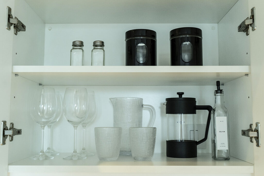 Interior de alacena mostrando copas de vino, vasos, jarra y prensa francesa de café, todo en cristal y blanco.