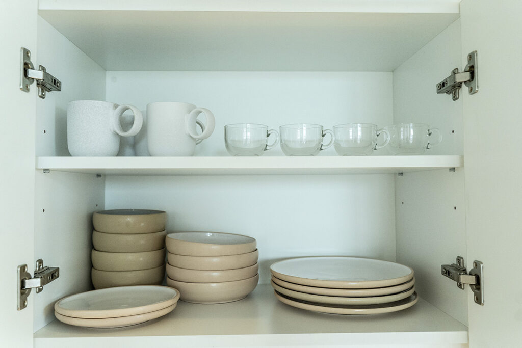 Interior de alacena con vajilla completa: tazas blancas en la repisa superior y platos en tonos beige y blanco.