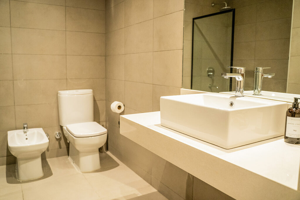 Baño moderno con inodoro, bidet, y bacha cuadrada sobre mesada, con espejo y toallero cromado.