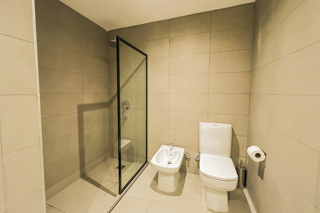 Baño completo con ducha de mampara de vidrio con marco negro, inodoro y bidet, con azulejos grandes en tonos neutros.